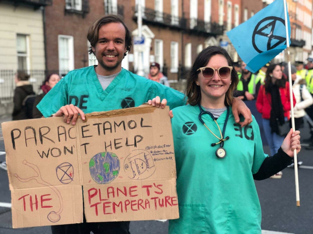 a male and female protestor in hospital gowns holding a poster reading "paracetamol won't help the planet's temperature"